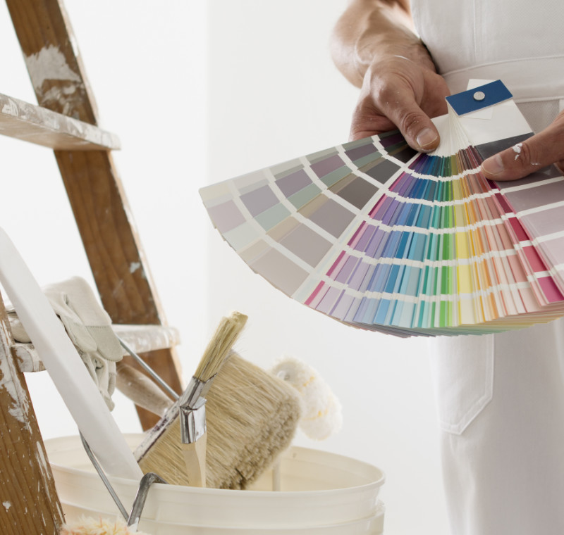 Hands Of House Painter Man Decorator Choose The Color Using The Sample Swatch, Work Of The House To Renovate, A Wooden Ladder With Paint Brushes And A Bucket As A Background, Close Up